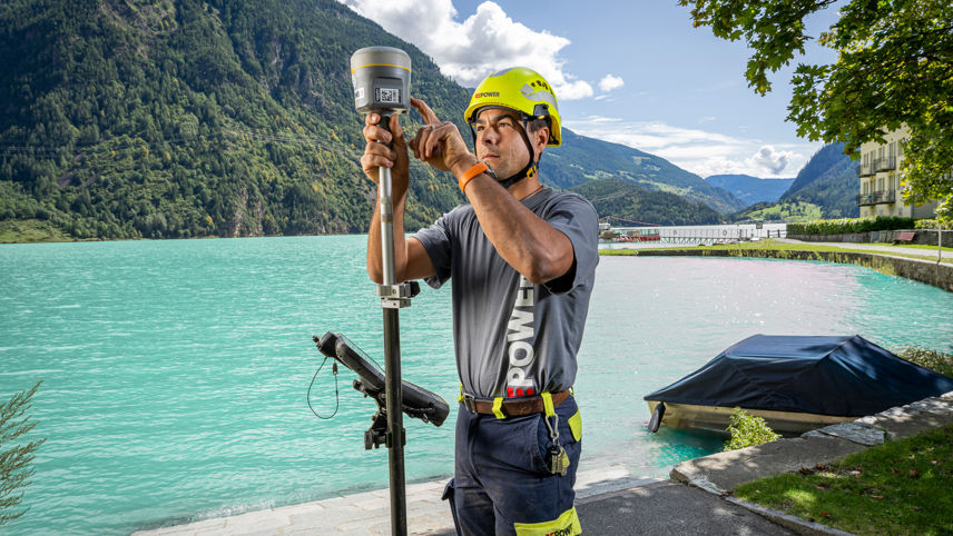 Technical specialist uses a GPS surveying device by a lake, with mountains and turquoise water behind.