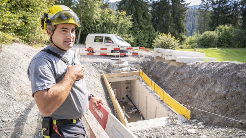 Grid electrician in a helmet stands at a construction site by a cable duct; barriers and materials behind.