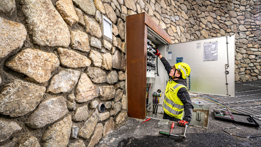 Grid electrician works on an open switch cabinet set in a stone wall; tools and cables are laid out.
