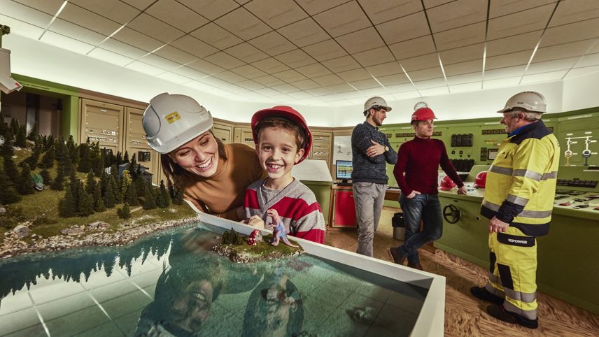 Guided tour at Cavaglia power plant: woman and child view a model; staff in the control room behind.